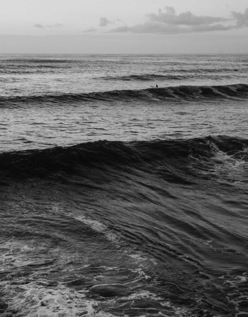 Black and white photograph of the ocean with a lone surfer on the horizon
